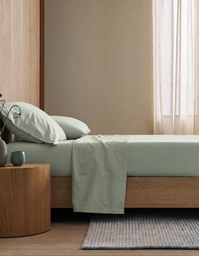 A minimalist bedroom features a wooden bed frame with Cozy Earth's Soft-Wash Cotton Flat Sheet in light green, two pillows, a round wooden nightstand with a green mug and vase, sheer beige curtains, and a light striped rug on wood floors.|Color:Laurel