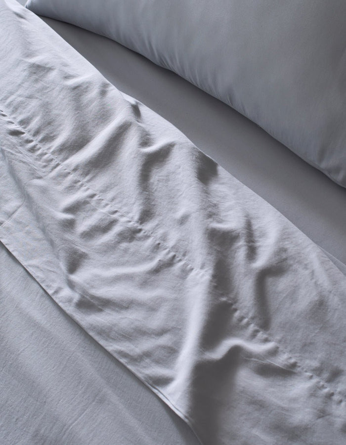 Close-up of a neatly made bed featuring Cozy Earth's Soft-Wash Cotton Sheet Set in light grey. Slight wrinkles add texture to the soft, clean fabric, giving the bedding an inviting look.|Color:Shore