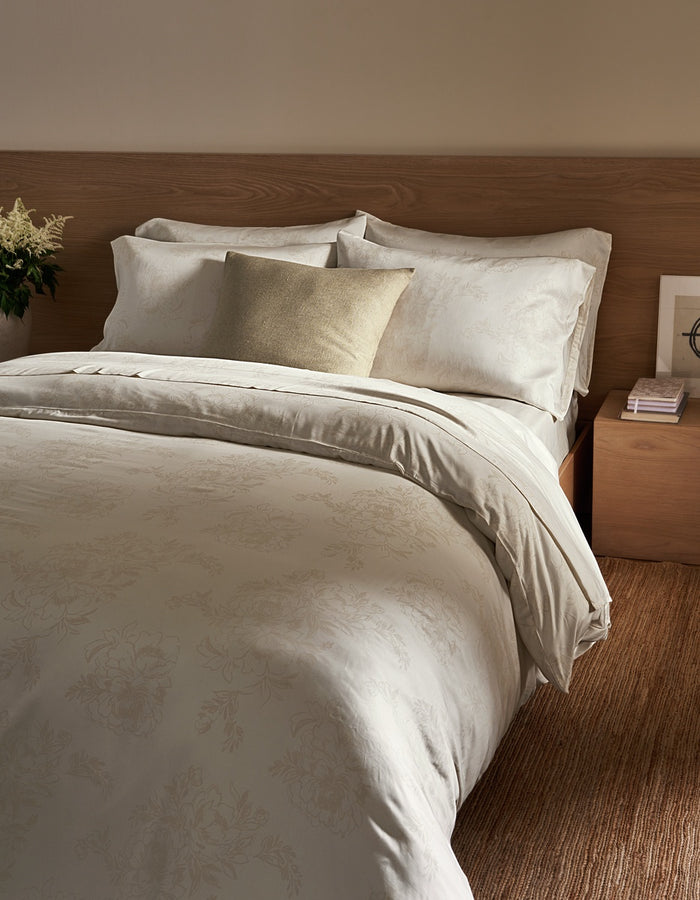 A neatly made bed with cream-colored bedding and a single Cozy Earth Sonoma Throw Pillow in beige, next to a wooden nightstand with stacked books and a small flower arrangement.|Color:Tallow|Size:24" x 24"