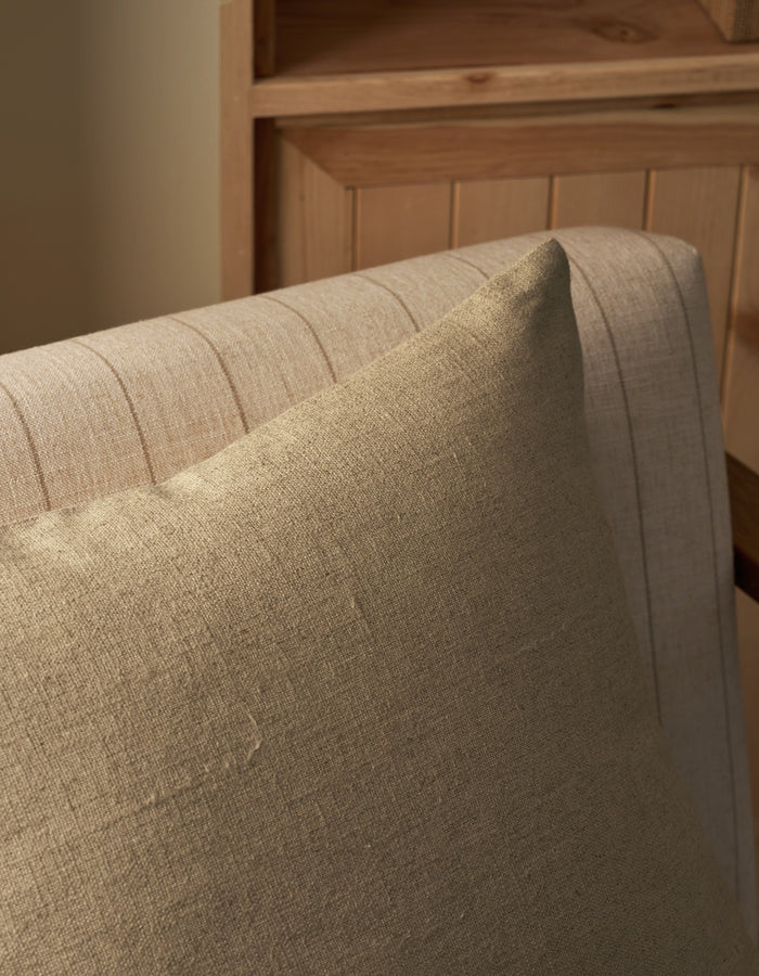 A close-up of the Sonoma Throw Pillow by Cozy Earth, in beige, resting on a light-colored striped upholstered chair, with wooden furniture in the background.|Color:Tallow|Size:20" x 20"