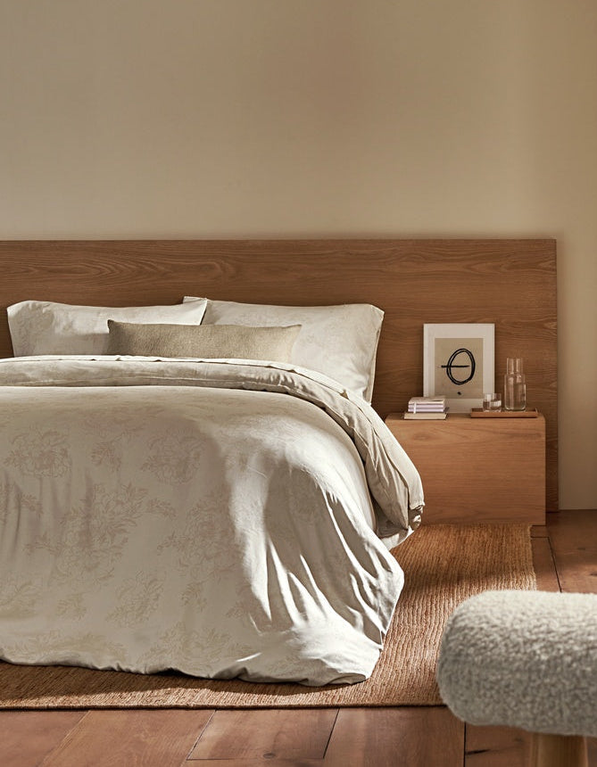 A cozy bedroom featuring a wooden bed with light bedding and accented by the Cozy Earth Sonoma Throw Pillow. A nightstand holds books and a glass of water, while sunlight streams through beige curtains on a large window. Wooden floors and shelves enhance the room’s warmth.|Color:Tallow|Size:14" x 30"