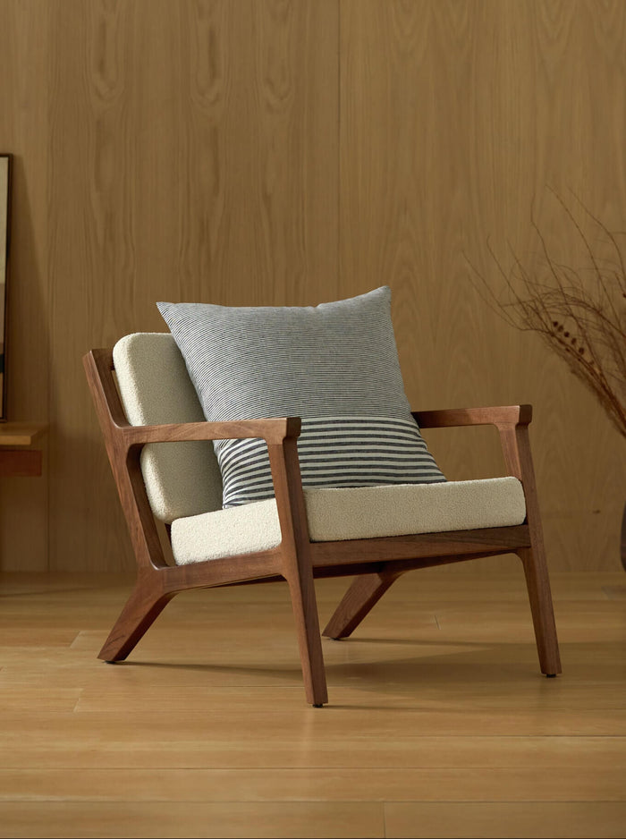 A modern wooden armchair with cream cushions and a Cozy Earth Striped Throw Pillow sits on a wooden floor before a light wood-paneled wall, with abstract art and a vase of dried branches in the background.|Color:Mixed Stripe Eclipse and Sea Salt |Size:24" x 24"