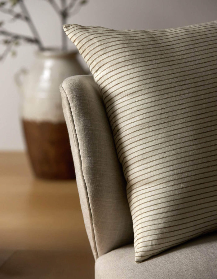 A close-up of the Cozy Earth Striped Throw Pillow resting on a light armchair, with a blurred ceramic vase and branches in the background.|Color:Narrow Stripe Sable and Coconut |Size:20" x 20"