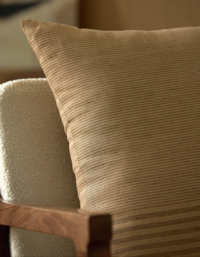 A close-up of the Cozy Earth Striped Throw Pillow resting against a light upholstered chair with a wooden frame. The softly blurred background creates a warm, inviting ambiance.|Color:Mixed Stripe Sable and Sandalwood |Size:26" x 26"