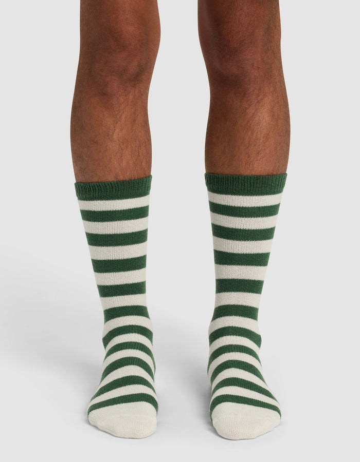 A person wearing The Striped Plush Lounge Sock by Cozy Earth, featuring knee-length horizontal green and white stripes, stands on a white background.|Color:Celadon Stripe