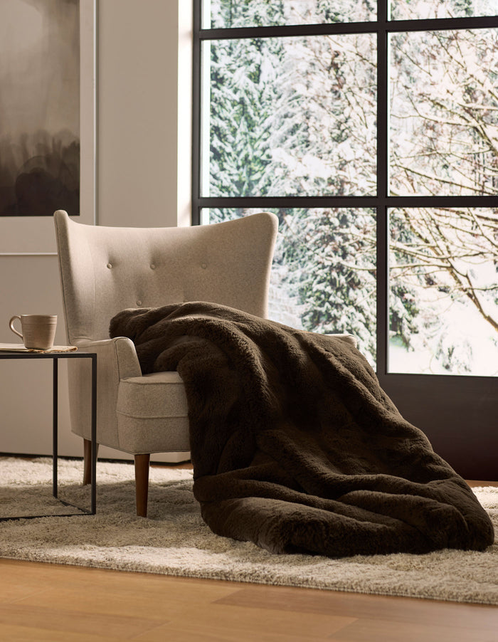 A cozy beige armchair with a Cozy Earth Cuddle Blanket draped over it sits next to a small table with a mug, overlooking snowy trees through large windows.|Color:Espresso