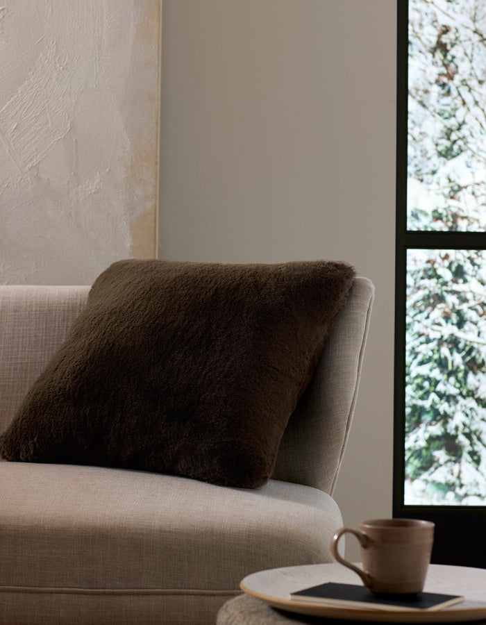 A beige sofa with the Cozy Earth Cuddle Pillow in dark brown sits by a window overlooking snow, while a ceramic cup and saucer rest on a small table nearby.|Color:Espresso |Size:24" x 24"