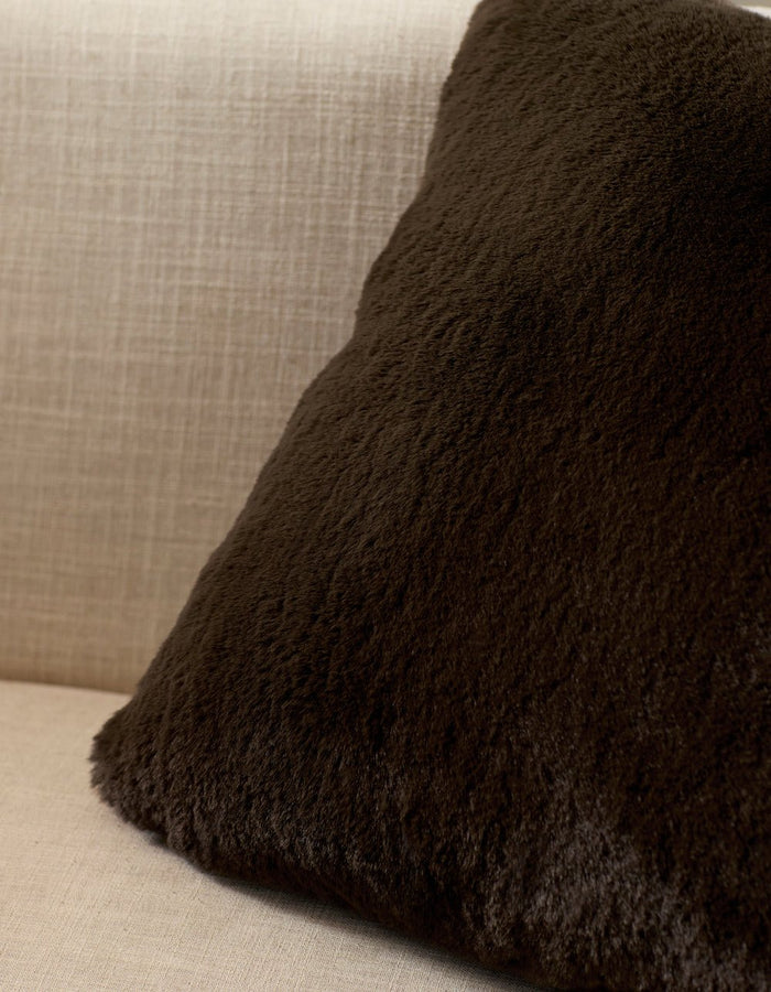 A Cozy Earth Cuddle Pillow in dark brown rests against the back of a light beige, textured fabric sofa.|Color:Espresso |Size:20" x 20"