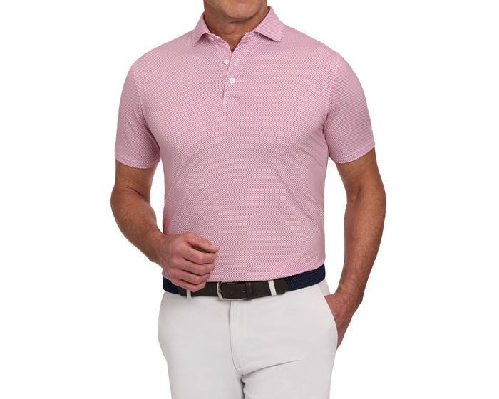 Man wearing The Armfield Shirt: Liberty Red, a pink geometric patterned polo with short sleeves, tucked into light trousers. Features a structured collar and woven belt.