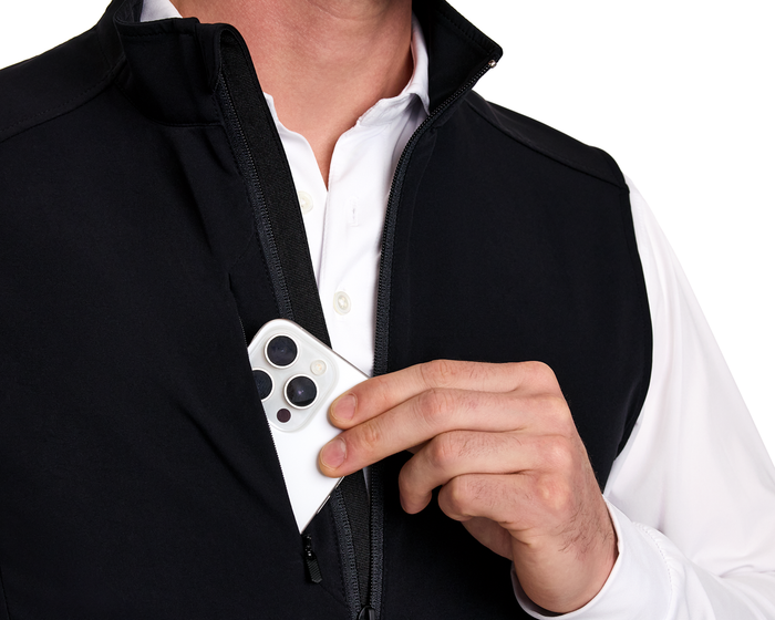 Person placing a white smartphone into the chest pocket of The Boyd Vest: Black, showcasing its sleek, windproof, and water-resistant design with a two-way zipper.