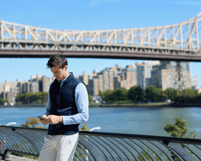 Man in The Cullinane Shirt: Heathered Vista Blue, leaning on railing by river, typing on smartphone. Urban skyline and bridge visible on a sunny day.