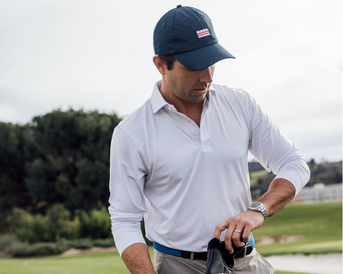 Male golfer wearing Holderness and Bourne men's white long sleeve polo and navy hat checks his watch on golf course.