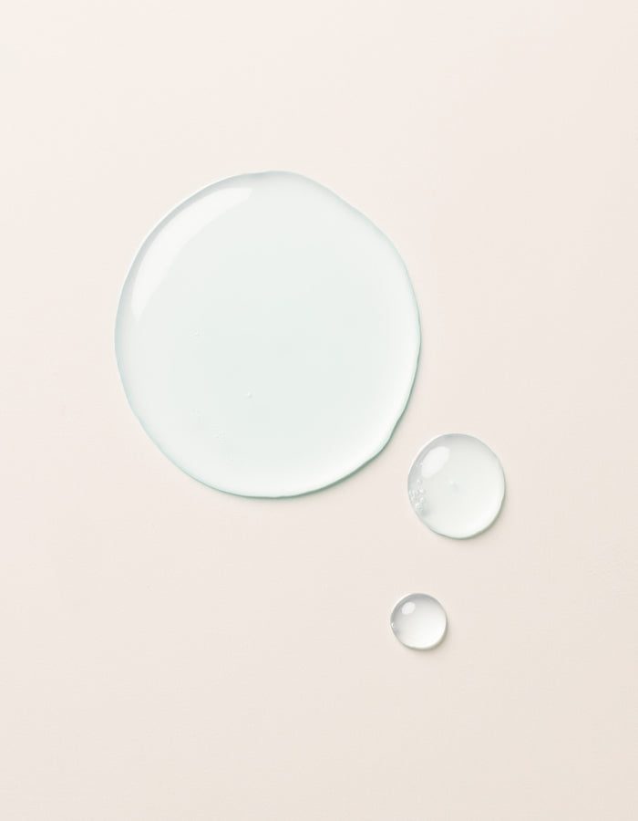 The Hydration Duo by Cozy Earth is shown as a large drop and two smaller drops of clear, light blue liquid on a smooth, light beige surface. The drops vary in size but all maintain a rounded shape, appearing glossy and slightly reflective.