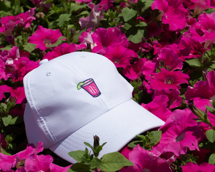 The Lightweight Performance Hat: White with Transfusion Icon, displayed among pink flowers, features a pink drink embroidery, adjustable fit, and UPF 50+ protection.