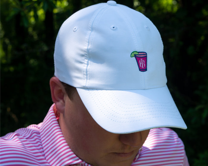 Person wearing The Lightweight Performance Hat: White with Transfusion Icon, featuring embroidered drink glass, paired with a striped polo, against a green blurred background.