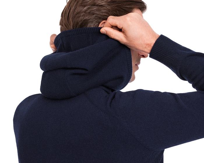 The Logan Hoodie Sweater: Heathered Navy, worn by a person pulling the hood, showcases ribbed detailing and a luxurious knit blend for a versatile look.