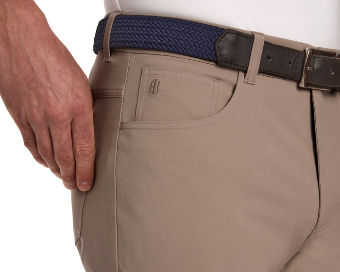 The McDaniel 5 Pocket Pant: Fescue, featuring light tan trousers with tonal logo, navy braided belt, and visible white shirt hem, showcases comfort and style.