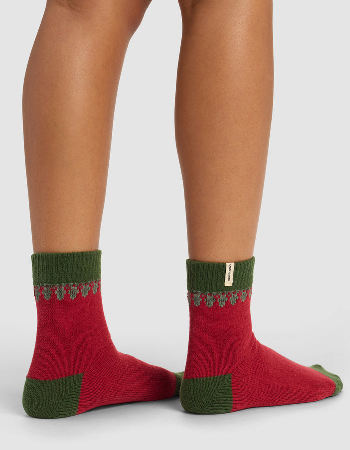 A close-up of a person’s lower legs in Cozy Earth’s The Mistletoe Plush Modern Crew Sock—red with green cuffs, heels, and toes, plus a small gray pattern near the cuffs—against a plain background.|Color:Cranberry