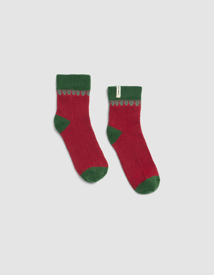 The Mistletoe Plush Modern Crew Sock by Cozy Earth features red fabric with green cuffs, heels, and toes, plus a decorative green and gray pattern on top. They are displayed flat against a plain white background.|Color:Cranberry