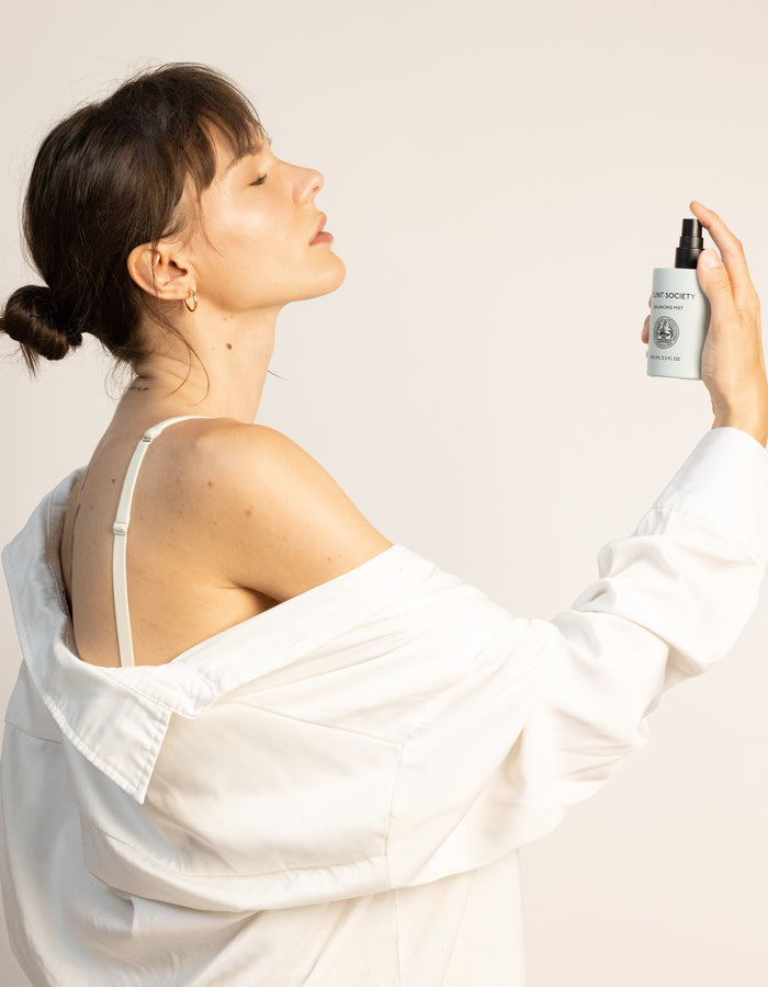 A woman with dark hair tied in a bun holds a bottle of Cozy Earth's Balancing Face Mist near her face, spraying it with a relaxed expression. She is wearing a white shirt that slips off one shoulder, revealing a white strap underneath. The background is plain and light-colored. |Size:Full Size