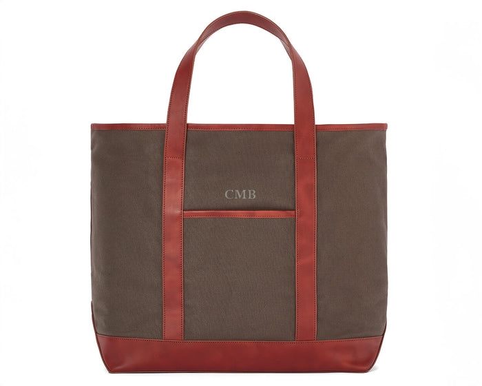 The Travis Tote Bag features brown canvas with reddish-brown leather trim, front pocket with CMB monogram, and is ideal for versatile use.