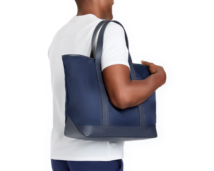The Treadwell Tote: Navy Ballistic - Slate Blue Embroidery, carried over the shoulder, showcases a sleek design with leather trim and functional interior pocket.