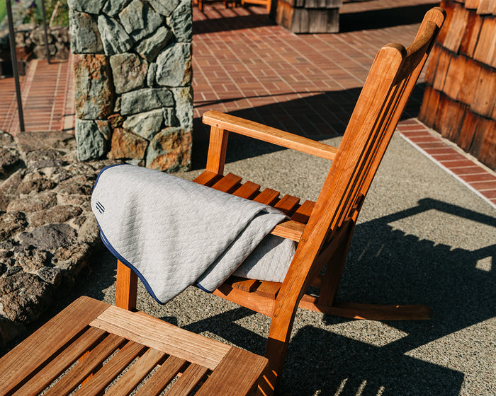 The Van Pelt Blanket: Heathered Gray, a cozy quilted blanket with dark trim, draped over a wooden rocking chair on a sunlit patio.
