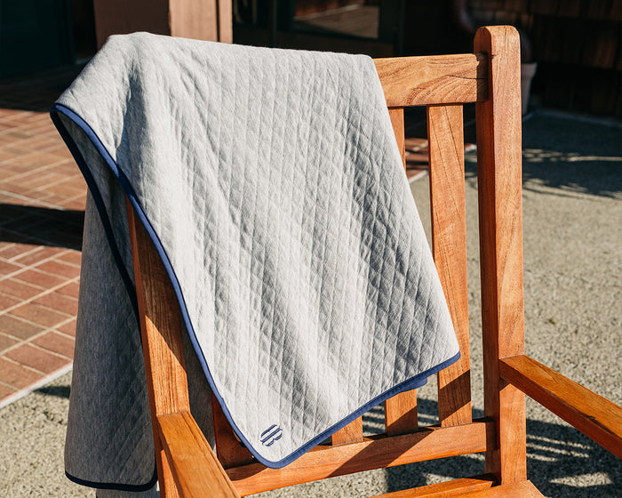 The Van Pelt Blanket: Heathered Gray, featuring a diamond-stitch pattern and dark-blue trim, draped over a wooden chair on a patio.