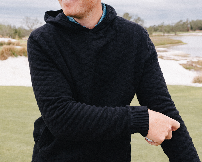 Person holding a golf ball, wearing The Wallace Hoodie Sweater: Black, showcasing its quilted design and ribbed panels for a modern, warm style.
