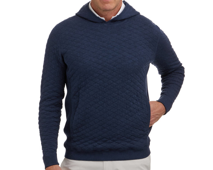 The Wallace Hoodie Sweater: Heathered Navy, a quilted pullover with ribbed cuffs, kangaroo pocket, and hood, worn by a person with hand in pocket.