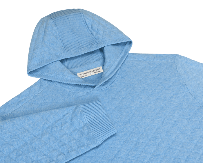 The Wallace Hoodie Sweater: Heathered Vista Blue features a light-blue knit pullover with diamond-quilted sleeves, ribbed hood, and cuffs, showcasing a streamlined, modern design.