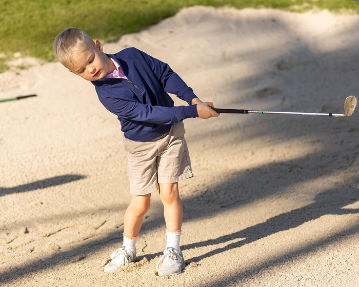 Young boy swings golf club in sand bunker, wearing The Westland Boys Quarter-Zip in navy, showcasing its performance fabric and versatile design.