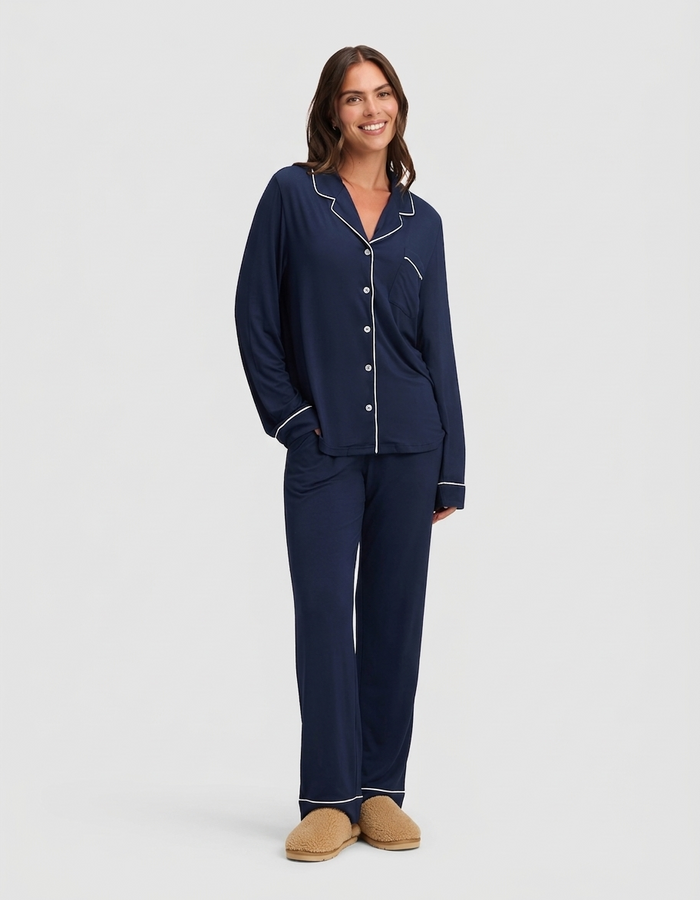 A woman smiles in Cozy Earth’s The Wind-Down Bundle, a navy blue pajama set with white piping, paired with beige slippers, against a plain light background.