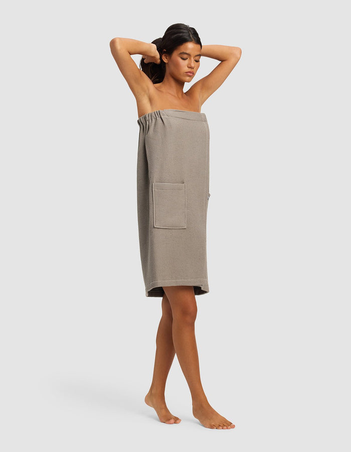 A woman with medium skin tone stands barefoot against a light gray background, adjusting her hair while wearing the Cozy Earth Waffle Bath Wrap in taupe, knee-length style with a front pocket.|Color:Charcoal