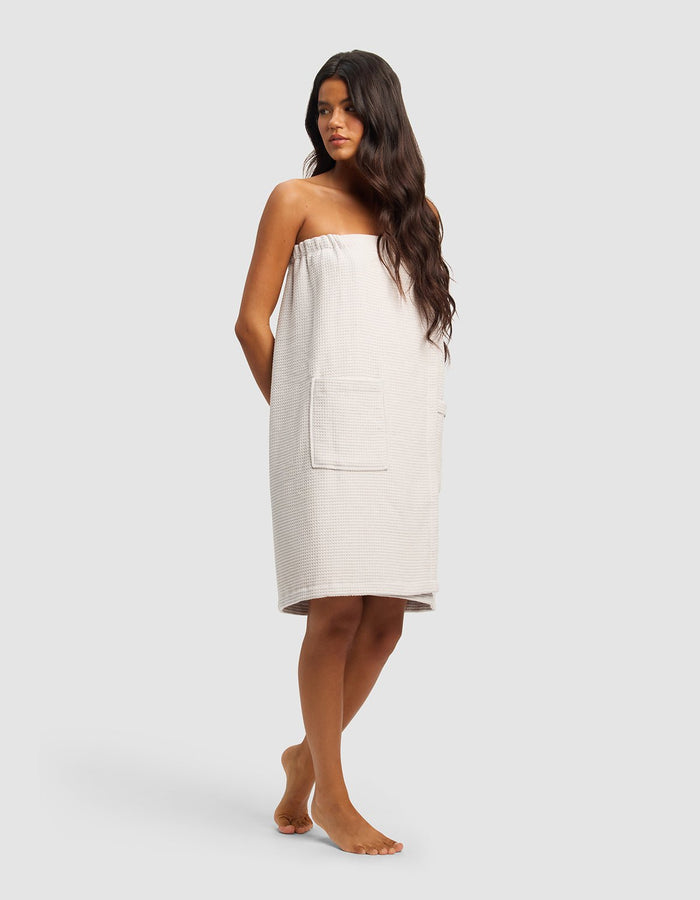 A woman with long dark hair stands barefoot, wearing the Cozy Earth Waffle Bath Wrap—a strapless, light towel wrap with large front pockets—facing slightly to the side against a plain, light background.|Color:Light Grey