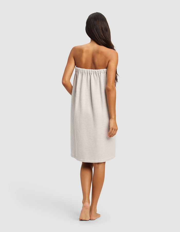 A woman with long dark hair stands barefoot, facing away, wearing the Cozy Earth Waffle Bath Wrap in light beige, covering her from chest to knees. The background is plain and light gray.|Color:Light Grey