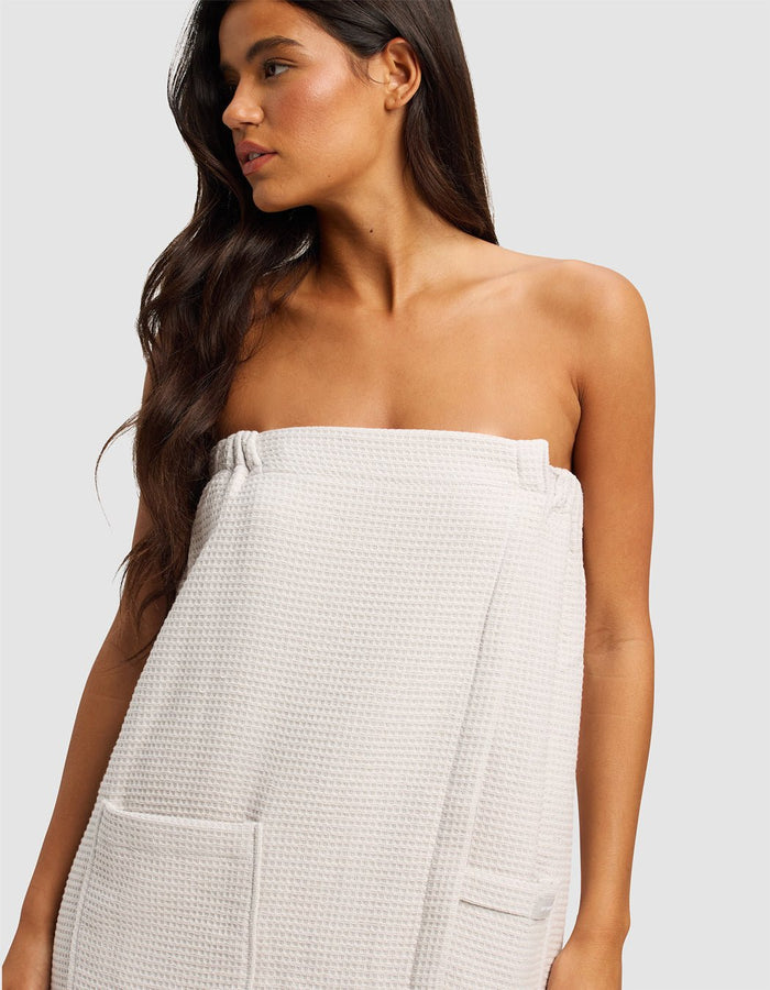 A woman with long, dark hair wears the Cozy Earth Waffle Bath Wrap, featuring pockets and a textured light fabric, standing against a plain light background and looking to the side.|Color:Light Grey