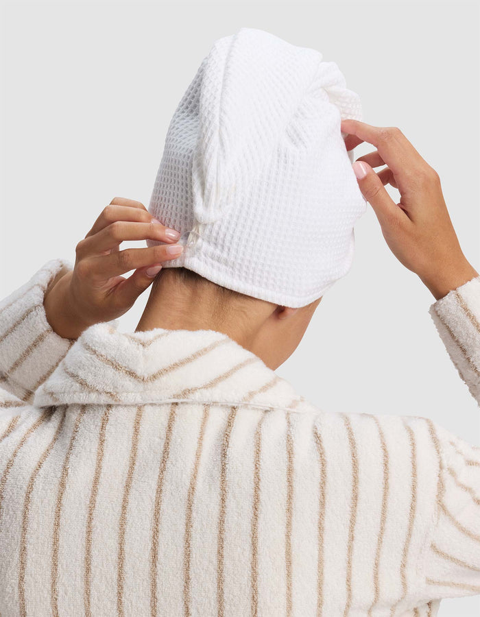 A person shown from the back, against a plain light background, wears a striped bathrobe and adjusts a Cozy Earth Waffle Hair Towel wrapped around their head.|Color:White
