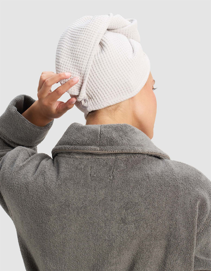 A person in a gray bathrobe is seen from behind, adjusting a Cozy Earth Waffle Hair Towel wrapped around their head. The background is plain and light-colored.|Color:Light Grey