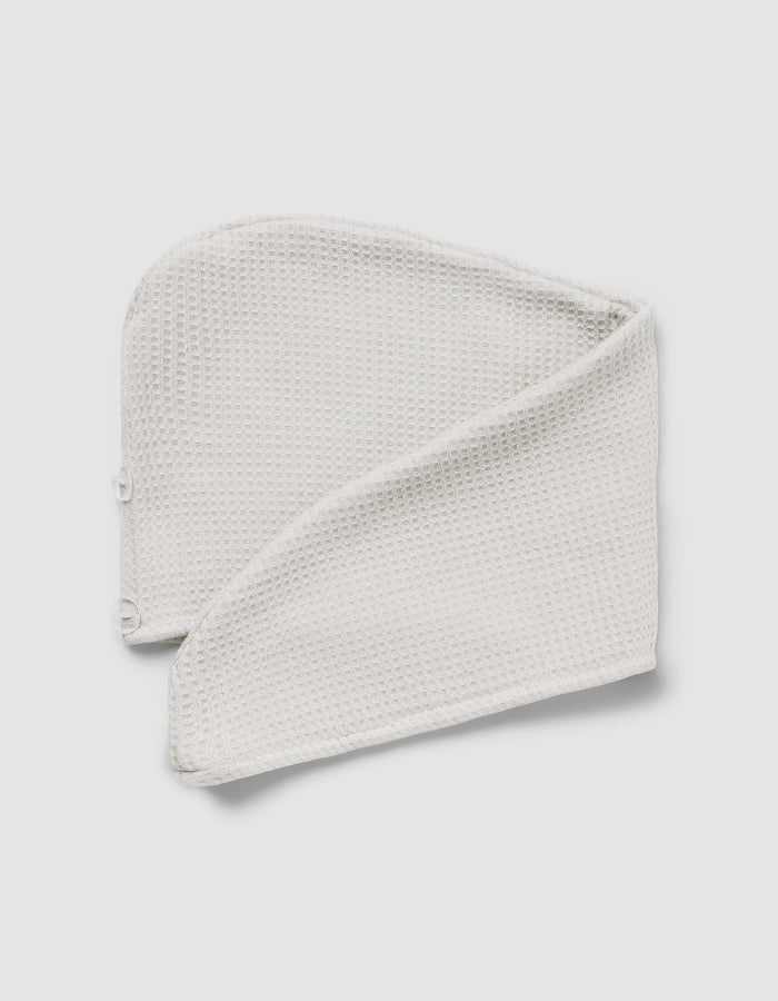 The Cozy Earth Waffle Hair Towel, a folded, textured white headband with snap buttons at one end, is displayed on a light gray background.|Color:Light Grey