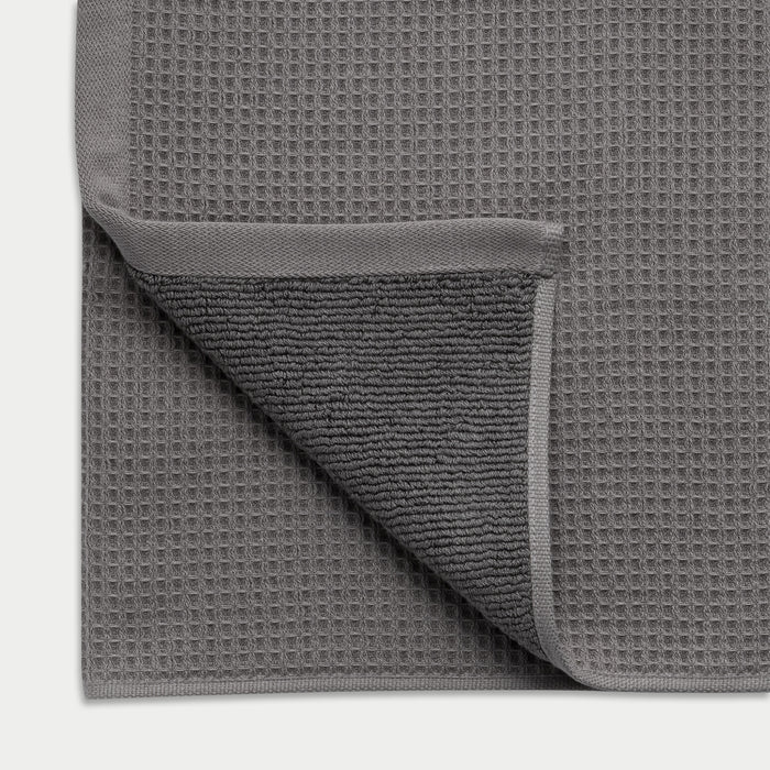 Charcoal Waffle Hand Towel resting flat on a white background. The corner is folded so that both sides of the towel can be seen. |Color:Charcoal