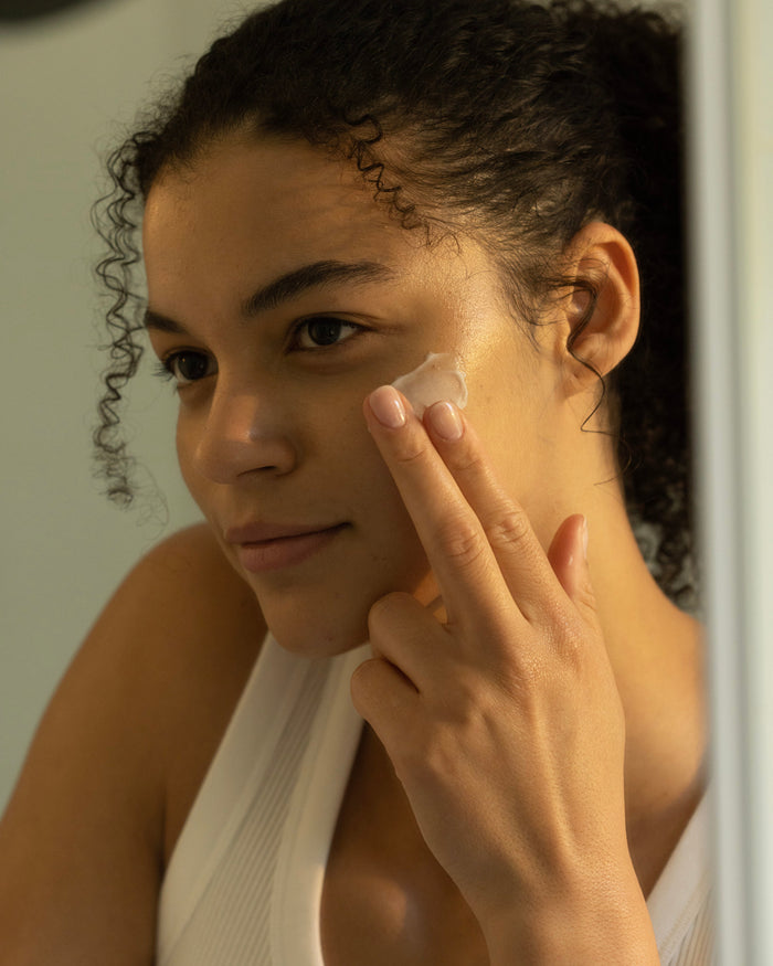 A person with curly hair is applying Cozy Earth's Water Cream Moisturizer with their fingers, concentrating on their left cheek. Their expression is calm and focused. They are wearing a sleeveless white top, and the background is softly blurred. |Size:Full Size