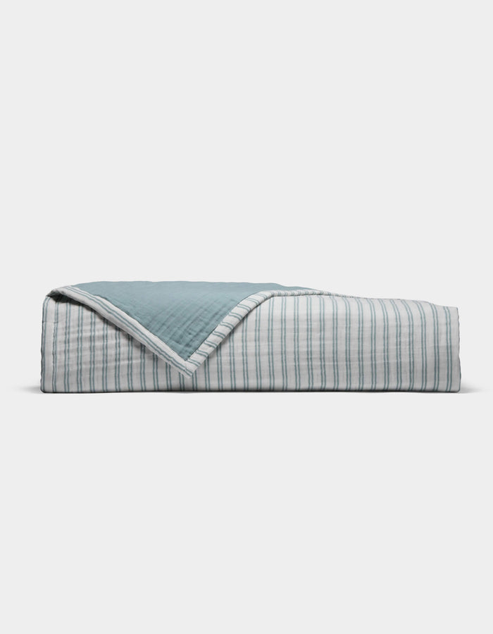 The Cozy Earth Waterfront Coverlet is a neatly folded quilt with a blue top and a white underside featuring thin blue stripes, displayed against a plain white background.|Color:River Stripe