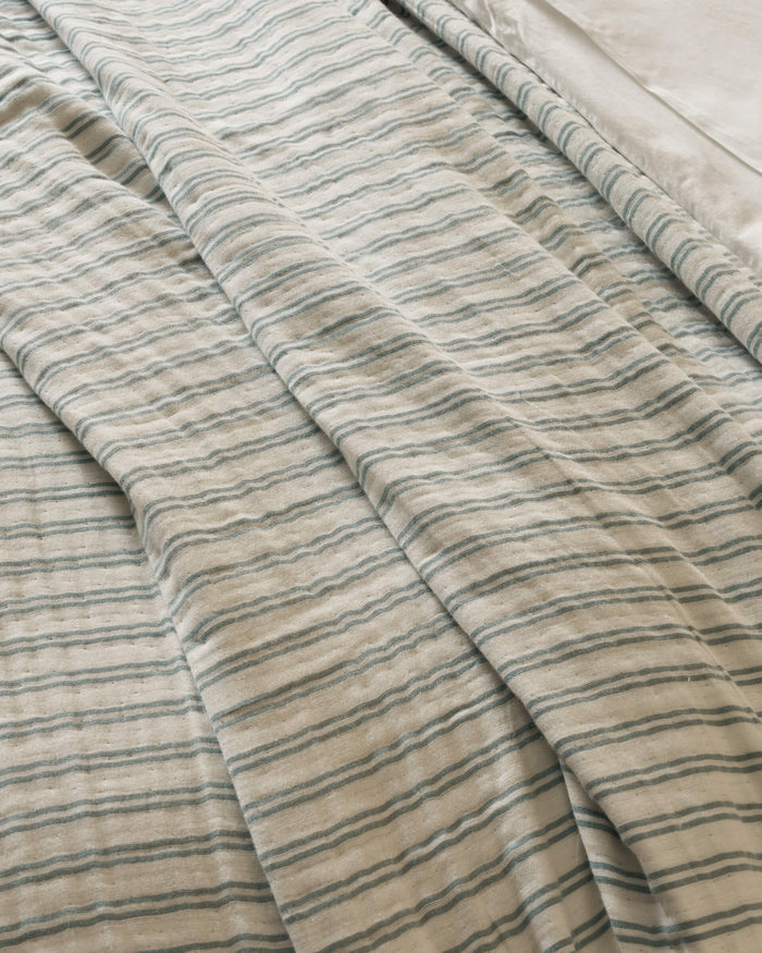 The Waterfront Coverlet by Cozy Earth is shown on a bed, displaying its cream color with thin blue-green stripes and gentle folds. A smooth white sheet is visible at the top right corner.|Color:River Stripe