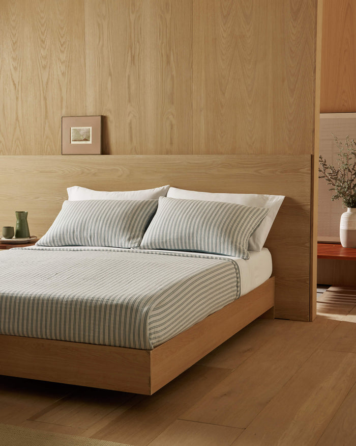 A minimal bedroom with light wood walls and floor, featuring a wooden bed styled with Cozy Earth’s Waterfront Coverlet, two pillows, a side table holding cups, and a vase with branches in the corner.|Color:River Stripe