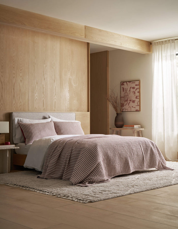 A modern bedroom featuring the Cozy Earth Waterfront Coverlet Bundle—pink-striped quilt and pillows—set against a light wood wall, with a beige rug, small side table, vase, framed abstract art, and sheer window curtains.|Color:Currant Stripe