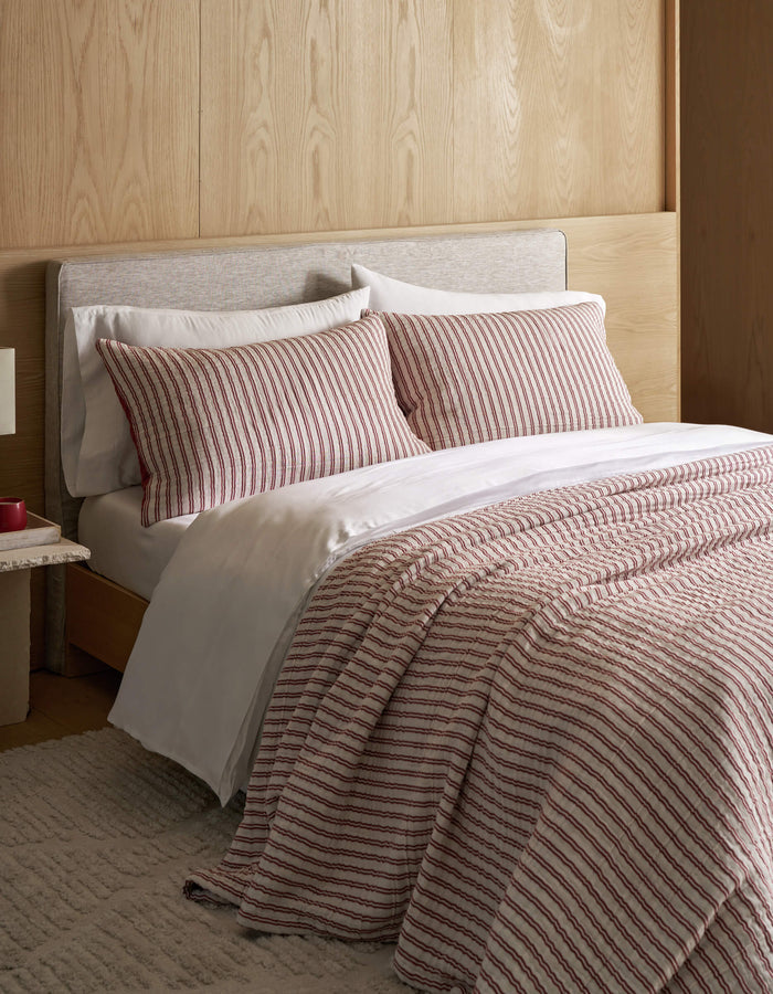 A neatly made bed features the Cozy Earth Waterfront Coverlet Bundle, white sheets, and a striped comforter with matching pillows, set against a light wood headboard and wall, complemented by a beige rug and a small bedside table.|Color:Currant Stripe