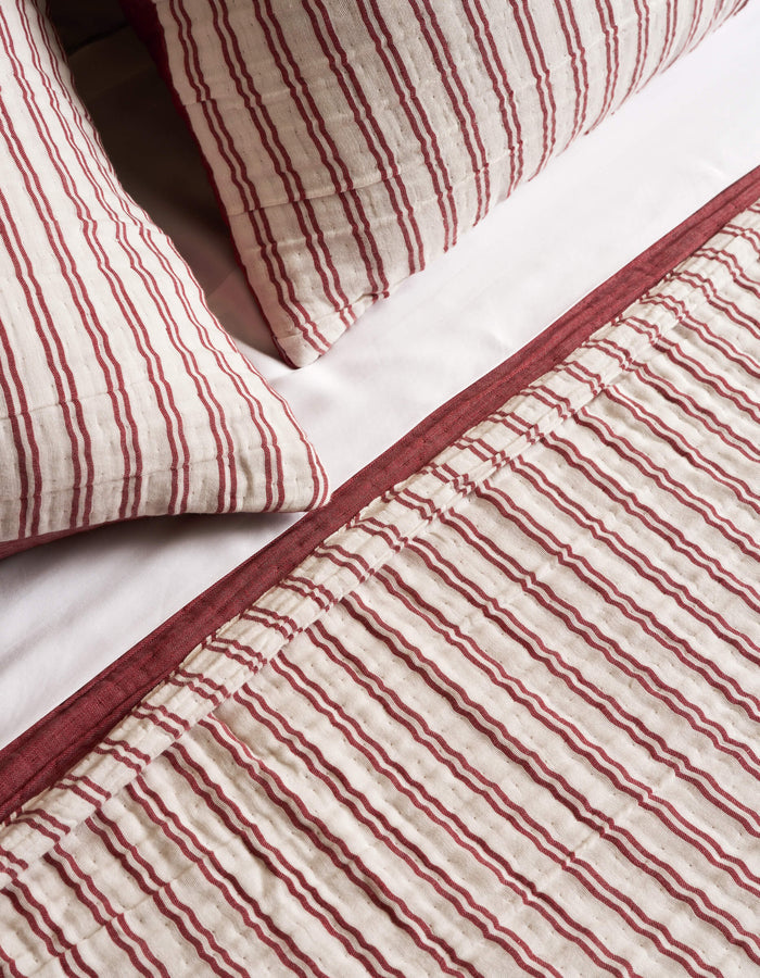 The Cozy Earth Waterfront Coverlet, featuring red and white vertical stripes with a solid red border, is shown on a bed with white sheets and two pillows.|Color:Currant Stripe
