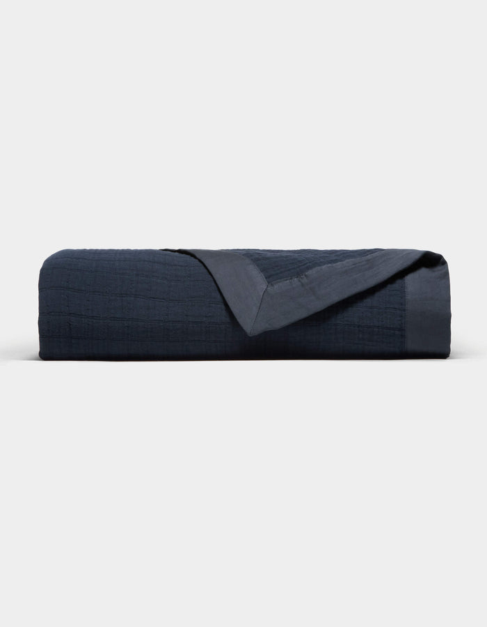 The Cozy Earth Waterfront Grid Matelasse Blanket is neatly folded in dark navy blue, showcasing its smooth, textured surface with one corner turned over, set against a plain light background.|Color:Eclipse