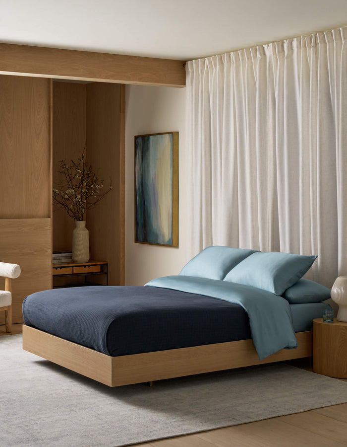 A modern bedroom features a light wood bed frame, the Waterfront Grid Matelasse Blanket by Cozy Earth in navy, light blue pillows, a large abstract painting, and a vase with branches on a wooden desk near floor-length white curtains.|Color:Eclipse